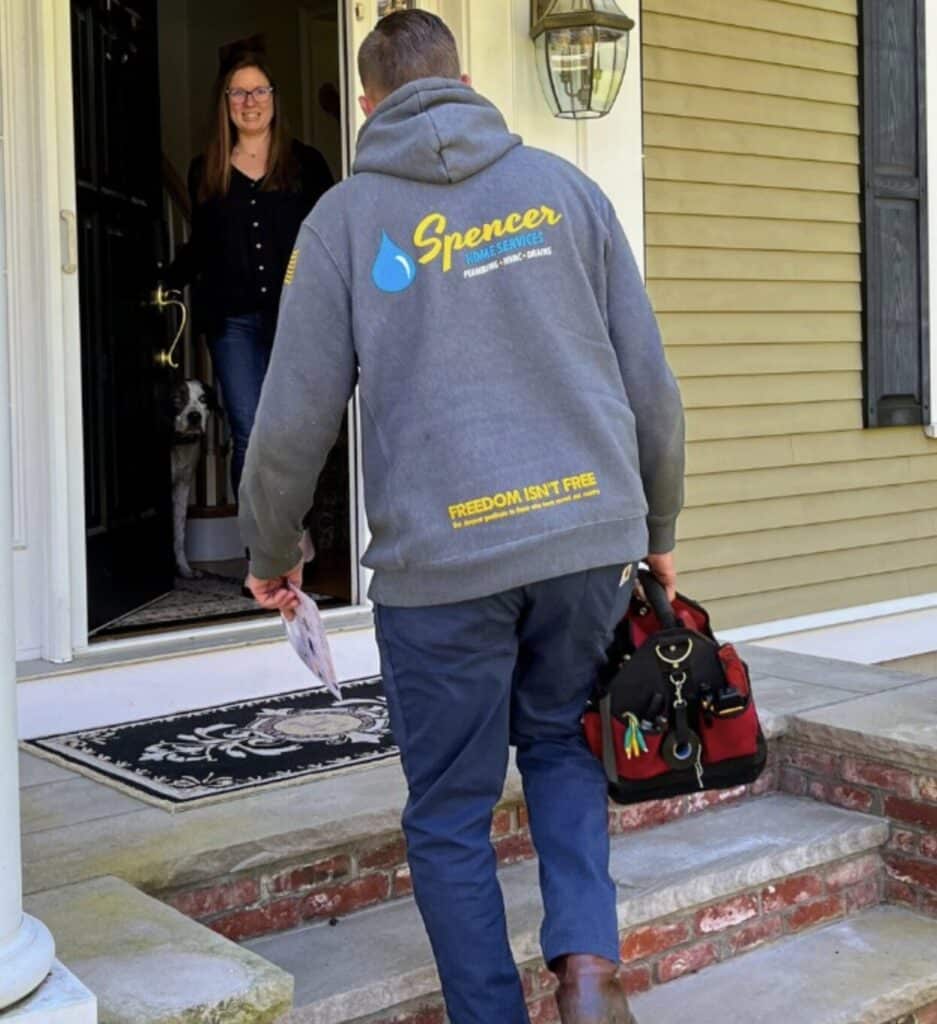 A Spencer Home Services technician carrying a tool bag walks up the front steps as a smiling homeowner and her dog greet him at the door