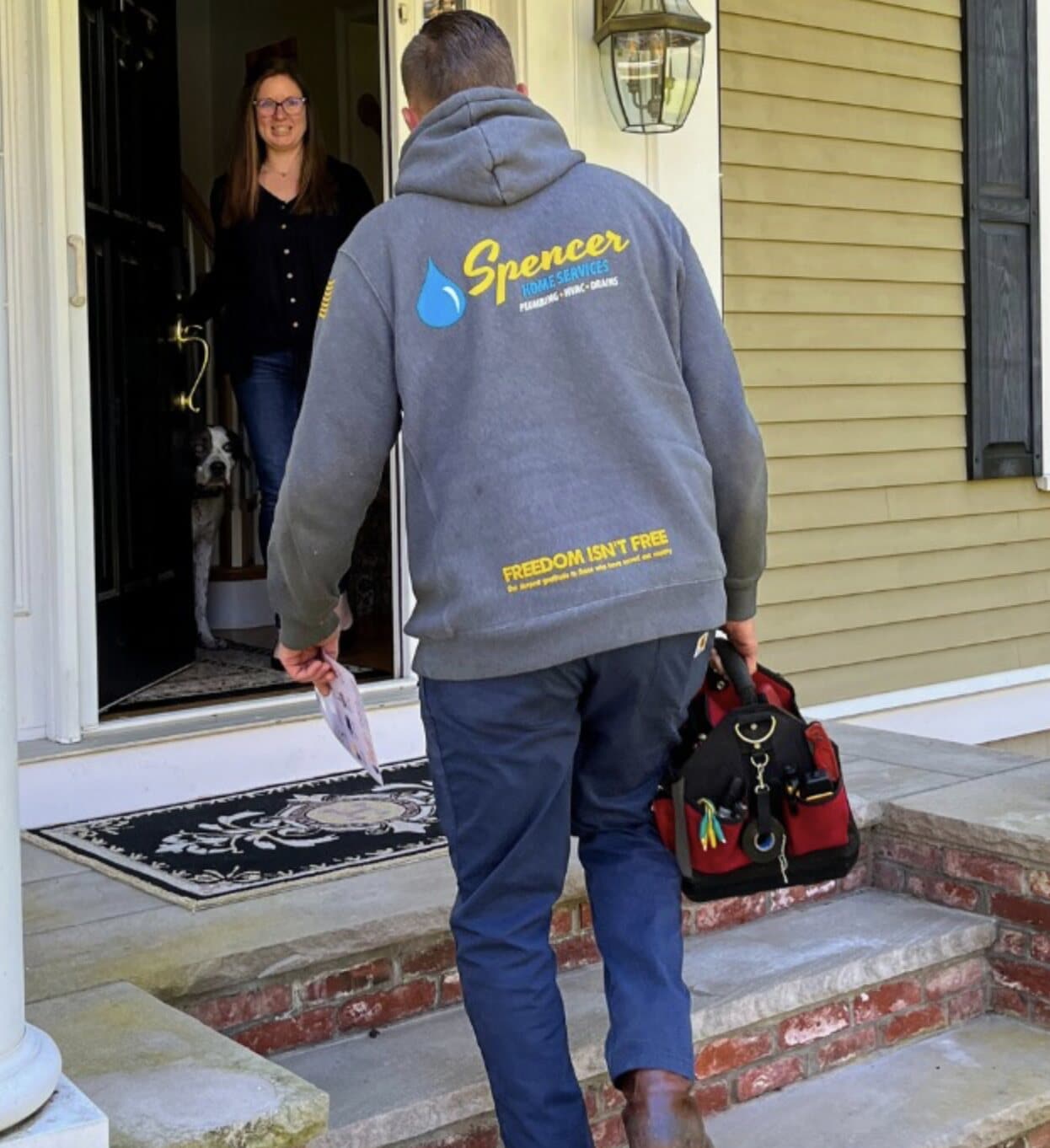 A Spencer Home Services technician carrying a tool bag walks up the front steps as a smiling homeowner and her dog greet him at the door